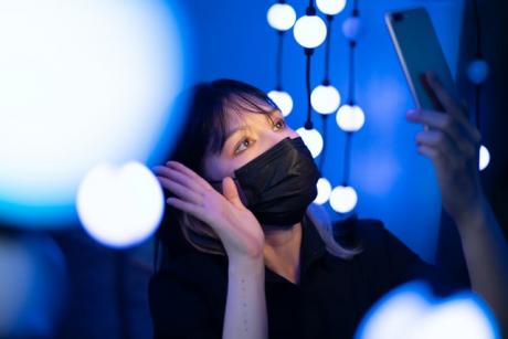 A woman wearing a mask uses her smart phone to video call at night surrounded by glowing blue lights. Bangkok, Thailand.